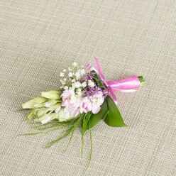 A single, elegant boutonniere is carefully laid on a softly textured, beige linen surface, the neutral fabric forming a quiet stage that lets every petal and ribbon detail shine. In the centre, a tight group of soft pink blossoms, their petals finely ruffled and slightly cupped, suggests miniature stock or similar cottage-garden flowers, gathered as if freshly picked from a border near Epping Forest. Their colour moves gently from a tender blush at the core to a lighter edge, giving depth without any harsh contrast. Threaded between these blooms are tiny sprays of white baby's breath, each minute floret like a star against the pastel tones, adding lightness and a nostalgic, bridal feeling often seen at summer weddings across Epping and nearby villages. Subtle hints of richer lavender or purple flowers sit just behind the main cluster, deepening the colour story while keeping the palette soft and romantic. On one side, a few slim, elongated flower buds in creamy white and pale green, much like freesia heads before they open, stretch towards the left, bringing a sense of movement and the promise of fragrance. Broad, glossy green leaves cradle the arrangement, their smooth surfaces catching the light, while finer, thread-like sprigs of greenery spill out delicately, echoing wild meadow grasses. Around the neatly trimmed stems, a bright fuchsia satin ribbon has been wrapped with care and tied into a clean, symmetrical bow, its sheen contrasting beautifully with the natural textures. The whole piece feels cool, fresh, and thoughtfully composed, the kind of handmade floral accent a local Epping groom or prom guest might choose to complement a tailored suit or evening dress. Crafted with the quiet precision you'd expect from an experienced florist, it captures the intimacy and celebration of a special day without ever seeming overstated.