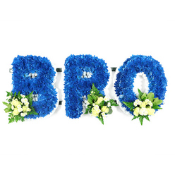 This reverent studio composition presents a three-piece floral tribute spelling 'BRO', each capital letter shaped from an abundant mass of royal blue chrysanthemum blooms that form a thick, plush surface. The arrangement is photographed against a stark white background, the clean negative space emphasizing the defined silhouettes of B, R and O and the thin white foam base that outlines their contours and supports the flowers. At the lower-left curve of the B and at the lower-right bases of the R and O are delicate accent groupings: pale cream to soft yellow spray roses in mixed opening stages, small white chrysanthemums or baby's breath for a whisper of texture, and a mix of glossy green foliage-broader salal or lemon leaf alongside lighter, feathery fern fronds. The colour interplay is intentional; the saturated blue conveys steadiness and remembrance while the creamy accents bring a soft, consoling light. The tactile contrast between the dense chrysanthemum heads and the airy sprays of filler invites a quiet reach to touch, and under soft, neutral lighting you can almost imagine the subtle scent of roses threaded through the clean, green aroma of foliage. As a local floral piece created for memorial use, this design reflects the understated sensibilities often requested for services in Epping and nearby Bell Common, and was carefully styled to lay respectfully at a chapel or graveside. Prepared in our Epping studio with traditional foam-mounted techniques, the tribute reads as both a personalised message and a composed statement of love and loss for a brother.