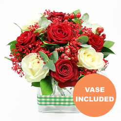 In this detailed floral image, a luxurious bouquet of red and white roses is presented front and centre, crafted with the kind of care you might expect from a local Epping florist who loves working with seasonal colours. The arrangement is dominated by plush, deep red roses and soft, creamy white roses, each flower fully open, with their layered petals forming graceful, cup-shaped blooms. Around them, clusters of tiny, vivid red filler flowers-similar to coloured gypsophila-create a fine, airy mist of colour, adding lightness to the dense rose heads. Interspersed red hypericum berries stand out as smooth, rounded beads, their lacquered surfaces hinting at winter berries found along paths near Epping Forest. Lush greenery anchors the display, with eucalyptus leaves and other mixed foliage adding tones of emerald and muted blue-green, giving a modern twist to a classic colour palette. The bouquet is arranged in a clear, rectangular glass vase, through which the neatly trimmed stems are visible, emphasising the tidy, professional finish. A green and white checkered gingham ribbon encircles the middle of the vase like a cheerful sash, suggesting a cosy, country kitchen or a festive table in a home near Epping High Street or Bell Common. In the lower right corner of the photograph, an orange circular graphic declares VASE INCLUDED in bold white capitals, making it clear that this floral gift arrives ready to place on a sideboard, office reception, or dining table. The overall mood is celebratory yet refined, perfect for anniversaries, winter gatherings, or a romantic surprise delivered across the Epping area.