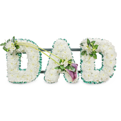 The image shows a thoughtful floral tribute spelling out "DAD" in bold, three-dimensional letters, designed as a memorial arrangement suitable for a funeral, wake, or cremation service in and around Epping. Each letter is completely filled with tightly clustered white chrysanthemums, creating a soft, cushion-like texture that suggests purity, peace, and quiet dignity. The flowers are fresh, with petals that look velvety and cool to the touch, and the uniform white surface is framed all around by a rich emerald green ribbon, gently ruffled to give depth and definition. Subtle glimpses of a dark supporting frame appear behind the letters, revealing that the tribute is mounted to stand upright, the way it might be displayed in a chapel of rest or along the aisle of a local Epping church. On the upper left arc of the first "D", a small spray of white freesia buds and lush green foliage adds a natural touch, as though freshly gathered from gardens near Epping High Street. Two slender green stems then sweep gracefully across from this area towards the central "A", guiding the eye to a more dramatic focal point. Here, among fresh greenery and more white freesia, sit two deep purple calla lilies, their sculpted, trumpet-shaped blooms introducing a solemn, jewel-toned accent to the otherwise white and green design. On the final "D", a smaller, coordinating cluster of white flowers and foliage is placed on the upper right curve, echoing the first spray without overpowering the overall balance. Set against a pure white background, the composition feels serene and deeply personal, a gentle way of saying goodbye to a father while surrounding his memory with calm, understated beauty.