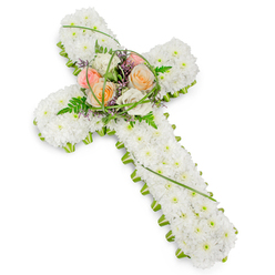 Set against a clean, white backdrop, this carefully crafted floral cross appears both substantial and tender, as if resting in still air. The cross itself is built from a generous bed of pristine white chrysanthemums, their small, rounded petals pressed closely together to form a soft, continuous blanket of bloom. This dense cushion of white flowers creates a sense of purity and calm, each floret contributing to a unified, peaceful surface. Along every edge of the cross, from the upper arms down to the base, runs a crisp trim of bright green foliage, offering a subtle yet decisive contrast that gently outlines the traditional Christian shape. At the heart of the cross, where the arms intersect, a slightly elevated cluster of larger flowers forms a focal spray. Here, roses in mellow shades of peach, delicate blush, and creamy ivory unfold their velvety petals, bringing a quiet warmth and human touch to the tribute. They sit among feathery fern fronds, slender arcs of vivid green grass, and small accents of dainty purple blooms that add depth and gentle movement to the design. The lighting is soft and even, with no harsh shadows, enhancing the sense of serenity and allowing the natural textures to speak for themselves. This floral cross would feel at home at a funeral or memorial gathering in Epping, perhaps placed beside the altar of a local church or in a chapel of rest near the High Street, offering a symbol of faith, remembrance, and lasting love during a time of loss.