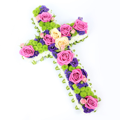 A cross-shaped floral tribute is displayed on a pristine white background, its colours and textures standing out with striking clarity, as if lit by soft daylight streaming into a quiet Epping chapel. The outline of the cross is crisply defined by a thick band of tiny white spray chrysanthemums, each floret pressed close to the next so the edge looks like a cushion of snow. Just beneath this white halo, a narrow band of folded pale-green ribbon or foliage wraps the perimeter, creating a tailored, almost garment-like finish that mirrors the fresh greens seen around Epping High Street in spring. Within this frame, the interior of the cross bursts with carefully balanced colour. Large roses in shades of vivid fuchsia and gentle cream or pale yellow open fully, their petals layered like silk, drawing attention to key points along the arms, centre, and base. Scattered between them are clusters of lime-green button chrysanthemums, their small, rounded heads providing a cheerful contrast that keeps the arrangement from feeling too heavy. Deep purple and violet blooms, likely lisianthus or statice, weave through the composition, adding shadows of colour and emotional depth, like quiet moments of reflection during a memorial service. The cross is photographed at a subtle diagonal, giving it a sense of movement and dimension, as if reaching outward in a gesture of comfort. This floral cross would be a fitting sympathy arrangement for a funeral or remembrance gathering in and around Epping, conveying peace, faith, and a gentle message of hope to grieving families.