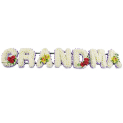 The image shows an elegant, bespoke funeral tribute spelling out GRANDMA in large, decorative block letters, arranged horizontally and photographed against a clean, bright white backdrop so that the floristry work appears crisp and uncluttered. Each letter is completely filled with tight clusters of small, snowy white chrysanthemums or similar button blooms, their petals overlapping to form a plush, almost cloud-like texture that suggests comfort and serenity. Around the base and edges of the letters, a fine border of soft lavender or muted purple flowers creates a subtle outline, giving the shapes definition and a hint of gentle colour when viewed from a distance, much as they would appear at a memorial service in Epping. On the G, a defined spray of three deep red roses, framed by glossy, dark green foliage, rests along the lower left arc, bringing a touch of rich, heartfelt emotion to the design. The first A is adorned with two striking chartreuse cymbidium orchids, their yellow-green petals and speckled reddish-brown centres adding a sophisticated, contemporary feel. In contrast, the letters R, N and D are kept purely in white and lavender tones, reinforcing the traditional symbolism of purity, peace and eternal rest. The M carries another trio of velvety red roses in its lower right section, visually balancing the arrangement, while the final A echoes the first with two more of the same yellow-green orchids, unifying the composition from end to end. All the letters are set onto a strong, unobtrusive dark frame that runs behind them, ensuring the tribute can be safely placed in a hearse, chapel, or at a graveside in and around Epping. The finished piece feels dignified and deeply affectionate, a tender floral expression of love for a much-loved grandmother.
