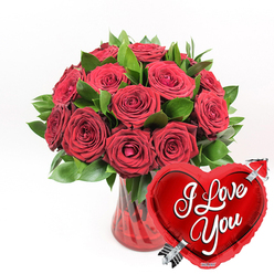 An opulent arrangement of red roses fills the frame, gathered in a clear ruby-tinted glass vase that seems to glow softly from within. The vase itself has a fluted, gently ribbed surface that catches and bends the light, giving the impression of quiet movement even though it stands perfectly still on the unseen surface. Around a dozen or slightly more roses have been carefully placed, each head angled so that their velvety petals can be appreciated from different viewpoints. Some blooms are wide open, revealing their spiralled hearts, while others are just beginning to unfurl, creating a rhythm of fullness and expectation. The rich, dark green foliage nestled between them feels lush and fresh, like leaves brushed with dew in the early morning around Epping Forest. The strong contrast between the saturated crimson petals and the cool green leaves gives the bouquet a dramatic yet timeless look. In the lower right, overlapping the vase, a glossy red heart-shaped foil balloon adds a playful and unmistakably romantic touch. Its surface reflects tiny highlights as though polished, and across its centre the message I Love You appears in flowing white script, clear and easy to read. A silvery metallic arrow, styled like Cupid's, appears to pierce the heart at a jaunty angle, making the sentiment feel both fun and deeply sincere. Presented against a flawless white background, the entire scene feels like a cherished Valentine's or anniversary gift ready to be delivered from a local Epping florist to a doorstep near the High Street, Bell Common, or a cosy home tucked off Theydon Road.