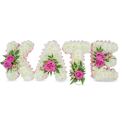 A sculpted, personalised floral display spells 'KATE' in substantial, three-dimensional block letters, each letter formed from hundreds of small, immaculate white chrysanthemums that create a seamless, cloud-like texture. A soft pale-pink satin ribbon neatly outlines the inner edges of every letter, giving definition and a whisper of blush against the white base. Bright hot-pink roses act as vivid focal points: a pair of opened pink roses on the K's left arm, a single prominent rose cradled low in the A, a lone vivid rose at the base of the T, and two roses accenting the right side of the E. Between these focal clusters sit delicate pink spray blooms, light alstroemeria highlights and varied greenery - ruscus, ferny fronds and variegated leaves - that lend contrast, depth and a natural garden freshness.

Presented freestanding on a pure white background with subtle shadows that emphasise its three-dimensional form, the tribute reads as both refined and heartfelt; the roses give a gentle, familiar scent while the chrysanthemums keep the overall tone calm and dignified. This elegant, personalised memorial piece is frequently chosen for funerals or celebrations of life in and around Epping, Station Road chapels and nearby Theydon Bois. Prepared with local care by Flowers Epping and available for delivery across Epping and surrounding villages via beville.live, it offers a tender, bespoke way to honour a loved one.