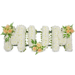 The image shows a refined funeral flower tribute that clearly spells out MUM in three bold, capital letters, all aligned in a straight line as if resting on a low stand. Each letter is densely packed with pristine white spray chrysanthemums, their small, rounded petals overlapping so closely that the surface looks like a soft, continuous cushion of bloom. Tiny shadows between the florets add subtle depth, while glimpses of dark green backing and ribbon edging trace the outlines, giving the letters structure and definition. To break the expanse of white and add a tender warmth, delicate accents of roses in soft peach and blush pink are arranged in three thoughtfully chosen places: at the top left of the first M, at the base of the U in the centre, and at the top right of the second M. These rose posies contain a mix of buds and half-open flowers, their spiral centres gently unfurling, surrounded by sprigs of rich green foliage and feathery ferns that curve naturally outwards. The whole composition is set against a clean, white background, which makes the colours and textures stand out without distraction. It feels like a tribute you might see at a quiet chapel in Epping, perhaps on the way to a service near the High Street or close to the edge of Epping Forest, where families gather to say goodbye. The design conveys calm, dignity, and gratitude for a mother's care, suggesting a message of peaceful rest and loving remembrance in every petal and leaf.
