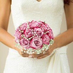 A hand-tied bouquet, central to an elegant Epping wedding moment, radiates intimate allure under soft natural light. Nestled in the crook of the bride's gently tanned arms, the bouquet's dozen pink roses display a painterly gradient-each bloom shifting from tender blush at the core to rich magenta edges, reminiscent of early blooms in the gardens near Epping's Market Square. Sprigs of baby's breath nod among the roses like silvery clouds, their delicate forms lending a sense of grace and nostalgia. The bride's slender fingers, finished with a transparent, glossy manicure, cradle the bouquet's stems just above a gathered sash, underscoring the effortless poise of bridal preparations along Lindsey Street. The white gown's intricate lace overlays the bodice with subtle floral motifs, echoing the bouquet's design and infusing the moment with a sense of tradition. Beyond, the neutral background suggests a timeless, serene setting, drawing focus to the careful artistry in every petal and ribbon. This image suggests the anticipation and gentle joy of a ceremony at Epping Forest's edge-a bouquet not just of flowers, but of cherished memories and new beginnings.