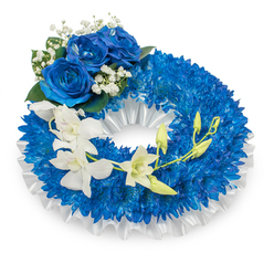 An elegant memorial wreath in a composed palette of sapphire blues and pure whites, presented on a clean white surface as if prepared for a chapel service on Epping High Street. The wreath's body is a sumptuous ring of densely clustered deep-blue chrysanthemums whose tiny, bristled petals create a tactile, cloud-like texture that absorbs soft, morning light. A ruffled white satin ribbon traces the inner opening, its gleam lending a serene frame against the vivid blue foundation. At the upper-left curve a small trio of striking blue roses opens fully, each petal soft and velveteen with a concentrated azure tone that complements the chrysanthemums while providing a focal point of reverent beauty. Circlets of delicate white Gypsophila nestle around these roses and lace through the blue mass, their whisper-thin stems and petite blooms adding an airy sensitivity. A graceful spray of white orchids - possibly Phalaenopsis or Dendrobium varieties - descends from mid-left toward the lower right, their porcelain-like petals and pale throats creating an almost floating movement across the wreath. Sprouting from the orchid stems are several pale green buds at varying stages, a subtle botanical symbol of ongoing life amid remembrance. Small touches of darker green foliage provide depth and natural contrast without overpowering the cool harmony. As a local florist I often prepare this style of tribute for families gathering near Epping Forest or at the station-side churches; Flowers Epping can deliver a similarly composed wreath for funerals and memorials across Epping and surrounding villages.