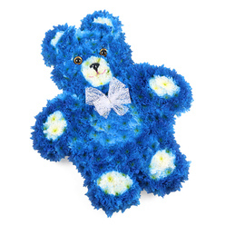 Set against a bright white backdrop, this three-dimensional floral teddy bear tribute appears almost like a beloved soft toy translated into petals, crafted with the kind of care you might expect from a long-established Epping florist. The bear's rounded body, head, and limbs are densely packed with vibrant cobalt and royal blue flowers, their small, layered petals forming an even, velvety surface that mimics plush fur. In contrast, soft cream and pale yellow blossoms highlight the key features: the inner cups of the rounded ears, the gently protruding snout, and the four oval pads on the hands and feet, each area clearly defined yet seamlessly blended into the blue. Two dark, round, glassy eyes catch the light, giving the bear a tender gaze, while a small black triangle nose and a simple curved line for a mouth suggest a quiet, reassuring smile. Around the neck, an airy white mesh bow, tied neatly with semi-transparent loops and tails, introduces a sense of celebration and innocence, like a gift prepared with love. The teddy lies flat, slightly angled within the frame, which emphasizes its cosy, resting posture and makes every detail of the floral work easy to see-the precise placement of stems, the uniform texture, and the fullness of the blooms. This design is particularly suited as a remembrance piece for a little one, a baby boy, or a child cherished by family and friends, adding a gentle, soothing presence to a funeral, chapel, or home in Epping, whether near the High Street, Stonards Hill, or the quieter residential lanes surrounding the town.