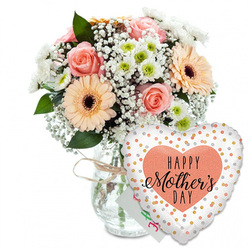 A tender Mother's Day scene unfolds against a clean white backdrop, where every stem and petal has space to breathe, much like the calm early mornings over Epping Forest. In the centre, a clear glass vase, shaped like a classic storage jar, holds a rounded bouquet arranged with the quiet confidence of an experienced local florist. Around the neck of the vase, a length of pale twine is tied in a relaxed bow, lending a touch of rustic charm that feels at home in Epping's mix of character cottages and modern houses. The flowers themselves are a gentle celebration of pastel colour: peach-pink roses fully open, their spiral hearts unfurled into soft, velvety layers, sit alongside large peach gerbera daisies with dark, almost chocolate-brown centres that draw the eye. Airy clusters of white baby's breath thread between the larger blooms, their tiny blossoms forming a soft halo, while compact white chrysanthemums with pale green centres add fullness and a crisp, garden-fresh brightness. Deep green foliage cradles the arrangement, creating contrast and a sense of lively growth, as if the bouquet had just been gathered from a well-tended Epping garden after a spring shower. Behind the flowers, a small, nearly hidden greeting card peeks out in white with hints of colourful lettering. In front, slightly to the right, floats a heart-shaped foil balloon: its white surface scattered with shiny gold and silver spots, framing a peach-coloured heart stamped with the words HAPPY Mother's DAY in bold black lettering and elegant script. The overall mood is one of affection and gratitude, perfectly suited for delivering love to mums across Epping, Loughton, and the surrounding area.
