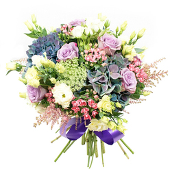 A lush tapestry of blooms takes centre stage, its artistry echoing the classic charm and natural beauty of Epping and its green surroundings. The eye is immediately drawn to the grand hydrangea heads, washed in cool hues of indigo, teal, and purple, much like the shifting shades seen in Epping Forest under changing skies. Nestled beside them, pale lavender and creamy white roses gently unfurl, their soft petals conjuring elegance with a hint of vintage allure-perfect for elevating events held in quaint local venues or gifting along Epping High Road. White ranunculus blossoms, tightly packed and layered, add a delicate texture and a sense of abundant fullness, while slim eustoma buds in pale yellow scatter brightness throughout the arrangement. Sprays of vivid fuchsia-pink filler and frothy pink astilbe create lively accents, almost as if echoing the wild, blooming corners of the nearby countryside. Fine strands of light green and lacy white filler flowers intertwine with glossy dark foliage, enriching the bouquet's depth and natural sophistication. Each element is expertly gathered and secured with a sumptuous, wide royal purple satin ribbon, harmonising the ensemble and adding a regal flourish that celebrates the area's refined taste. Whether shared to mark a milestone or to bring a sense of serenity into a home overlooking Epping's rolling fields, this professionally crafted bouquet speaks to heartfelt celebrations and the enduring beauty of local flowers, freshly arranged and ready for delivery across the community.
