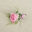 A single blush pink rose boutonniere is carefully styled on a softly textured light beige linen cloth, the kind of natural fabric you might see on a rustic wedding table in Epping High Street restaurants. The rose is semi-open, its velvety petals spiralling out from a gentle pastel heart to deeper pink edges that curl slightly, suggesting a bloom cut at perfect freshness that would give off a subtle, sweet fragrance. Clusters of creamy white baby's breath nestle tightly around the base of the rose, their tiny, cloud-like flowers creating a delicate halo and soft contrast against the richer pink. From behind the rose, fine, grass-like blades of emerald green foliage fan outward in graceful arcs, adding airy movement and framing the central flower without overwhelming it. A larger, darker green leaf is folded neatly around the stems, cradling the rose and baby's breath like a tailored collar on a groom's suit. The stems are wrapped in a lustrous satin ribbon that blends lavender-pink with deeper magenta-purple tones, the smooth fabric catching the light and adding a refined, romantic accent. The knot is simple and elegant, echoing the understated sophistication of a classic Epping church or registry office ceremony. Soft, even lighting creates gentle shadows along the woven linen and around the petals, giving the image depth while keeping the focus on the intricate craftsmanship. This boutonniere feels ideal for a groom or prom date heading out through Epping Forest Gate or past St John's Road, carrying a touch of timeless romance on their lapel.