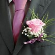 There's a lyrical beauty to this buttonhole arrangement, much like the gentle energy that radiates through Epping's village greens on bright mornings. At centre stage nestles a pink rose, its velvet petals softly unfolding to reveal a fresh blush that suggests affection, hope, and the excitement of a big day ahead. Around this central flower, trails of baby's breath lightly frame the rose, the clusters of tiny, papery white blooms lending an almost whimsical, cloud-like accent. Wisps of slender greenery and broad, glossy leaves stretch elegantly behind, adding movement and a sense of woodland freshness drawn from the outskirts of Epping Forest. The ensemble is finished with a lavender satin ribbon, tied gracefully and shimmering under soft natural light; it echoes the delicate tones of the rose while adding modern elegance. Anchored to the lapel of a charcoal grey suit-its fabric textured and pristine, complemented by a bold fuchsia tie and a subtle, patterned grey shirt-the boutonniere glows as the signature detail for someone celebrating love or achievement in the heart of Epping. This creation, meticulously crafted by a local florist, feels like a poetic tribute to milestones, infusing each moment with quiet anticipation, joy, and the warmth unique to the Epping community.
