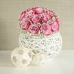 A softly lit indoor scene shows a lush, rounded bouquet of around fifteen to twenty soft pink roses, each bloom opening generously so the petals fade from a pale blush at the centre to a deeper, almost fuchsia pink along the ruffled outer edges. Between the roses, tiny white baby's breath flowers create a misty, cloud-like veil, giving the arrangement an airy lightness and a romantic, almost bridal feel. The bouquet sits snugly in a spherical white vase woven from natural fibres, its open, criss-cross pattern reminiscent of painted wicker or rattan, so that small pockets of shadow and light play through the gaps. This textured vase rests on a pristine white surface, as smooth as a freshly polished table in a calm Epping living room, subtly reflecting the gentle daylight that seems to filter in from a nearby window. To the left of the vase, two decorative spheres made from light beige, interlaced wooden strips add balance and interest; one is noticeably larger, both echoing the natural, handcrafted style. The background is a warm, softly textured beige wall, unobtrusive yet cosy, like the neutral tones often found in homes around Bell Common or along Epping High Street. The overall mood is serene, fresh, and quietly sophisticated, suggesting a thoughtfully styled corner for a bedroom, hallway console, or intimate dining table. It feels perfect for celebrating a tender anniversary, Mother's Day breakfast, or a gentle thank-you, capturing that blend of modern simplicity and heartfelt warmth that so many people in Epping favour for their everyday flowers and special occasion décor.