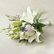 A lush, contemporary bridal bouquet from an Epping florist is artfully arranged on a pale, woven linen, evoking the tasteful interiors of local homes and wedding venues. The bouquet's centrepiece is a collection of show-stopping white Oriental lilies-some in full bloom, others delicately unfurling-each a symbol of purity and new beginnings. Their golden anthers and subtly speckled throats add gentle contrasts. Nestled throughout are smaller clusters of Lisianthus, ruffled and edged in vibrant purple, as well as a few tightly closed buds waiting to open. Sprays of dainty purple Veronica and airy white Gypsophila introduce depth and a dreamy softness, while layered green leaves and feathery fern provide a rich, natural foundation. The floral stems are elegantly gathered and secured with white ribbon, reflecting the attention to detail and artistry found in Epping's trusted flower shops. Washed in soft, diffused light, the bouquet's cool whites and vivid purples appear radiant against the understated background. The overall effect is one of sophisticated serenity, perfectly suited to a wedding at Epping Forest or as an anniversary gift-an arrangement that feels both fresh and timeless, crafted with the care and knowledge of a true local expert.