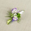 A finely crafted wedding boutonniere rests against a softly textured light beige linen backdrop, the neutral fabric allowing every detail of the flowers to stand out with quiet elegance. At the heart of the design are two open lisianthus blooms, their petals a clean, luminous white with carefully ruffled edges brushed in deep, royal purple, creating a rich halo of colour around each flower. Nestled between them sit tiny white filler flowers, similar to baby's breath, forming a cloud-like softness that lightens the composition. A slender vertical stem of muscari, with tightly packed, bell-shaped florets in a saturated grape-purple tone, adds height and a slightly wild, garden-picked charm, reminiscent of spring mornings around Epping Forest. Pale green, unopened buds tuck in close to the main flowers, suggesting freshness and the promise of new beginnings, a fitting symbol for a wedding or prom held in and around Epping's churches, barns, or registry office. Fine, feathery greenery, likely asparagus fern, fans out delicately behind the blooms, its airy texture softening the edges and giving the buttonhole a natural, freshly gathered feel. Broader, darker leaves provide a subtle framework, helping the white and purple tones to glow against the background. The stems are neatly gathered and wrapped in a crisp white satin ribbon, smoothed and secured with a tiny, precise bow, ready to be pinned to a lapel without fuss. The overall mood is refined yet gentle, like a thoughtful finishing touch for a groom or groomsman walking through Epping High Street on the way to a heartfelt celebration, the flowers adding a whisper of colour and scent rather than a shout.