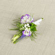 A considered study in soft contrasts, this boutonniere pairs two white lisianthus edged in violet with frothy sprigs of baby's breath and a playful pulse of grape hyacinth, all tied into a compact, wearable work of art. The lisianthus sit like two open promises, their silk-fine petals cooling to the touch and edged with a delicate lavender rim that reads both classic and modern. Around them, clusters of tiny white filler flowers create a gentle, clouded softness while slim buds and the upright, cobalt-tinged spikes of grape hyacinth introduce a liveliness that nods to spring's arrival. Wispy ferns reach out behind the flowers, their textured fronds offering an almost woodland whisper that balances the arrangement's polite formality. The stems are elegantly bound with white satin ribbon, finished in a neat knot that will sit snugly on a lapel without bulk. Placed on neutral linen, the colors hold their quiet energy: cool whites, tender violets, and a clarifying blue-violet that reads as both refined and joyous. As a florist working from Flowers Epping near the town centre, I assemble boutonnieres like this for couples heading to small local venues and gardens around Theydon Bois and Bell Common; the design is chosen not only for its visual effect but for the sensation when worn - the gentle weight, the whisper of ribbon, and the subtle floral scent that accompanies signatures and photographs. It's a small piece that carries a lot of intention: thoughtful, restrained and utterly suitable for weddings, commemorative portraits, or milestone ceremonies held close to home.