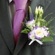 A close-up photograph captures the upper chest of a groom or groomsman dressed for a wedding in Epping, the focus resting on a finely tailored dark charcoal grey suit jacket and its elegant boutonniere. The lapel is sharp and structured, lying over a pale grey checkered dress shirt whose fine pattern adds quiet texture beneath a lustrous magenta silk tie, knotted with precise care and glowing softly in the light, much like you might see at a ceremony near Epping Forest. Pinned to the left lapel, the buttonhole is a small work of floral art: two white lisianthus blooms edged dramatically in rich purple, their ruffled petals cupping sunny yellow centers that seem to radiate warmth. Around them, slim unopened white buds reach upwards, suggesting blossoms still to come, while a slender spike of deep indigo-purple veronica rises to give vertical elegance and a slightly wild, garden-grown feel. Tiny white filler flowers, similar to baby's breath, tuck in and out of the composition, softening the edges and lending a bridal delicacy. A mixture of fresh greenery frames the flowers-fine feathery fronds, smooth emerald leaves, and loops of decorative green wire or grass that echo modern wedding styling often chosen by local couples along Epping High Street. At the base, a neat white satin ribbon bow secures the stems, its gentle sheen catching the light. The overall impression is one of polished sophistication with a romantic heart, a single floral accent designed to sit close to the wearer's heart during vows, photographs, and celebrations that will be remembered for years.