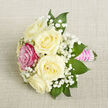 On a softly textured, light beige fabric backdrop, a small hand-crafted bouquet lies on the diagonal, as if just set down before a special moment begins. The eye is first drawn to a cluster of creamy white roses, five in total, each bloom fully formed yet not overblown, their petals curling outward in smooth, rounded layers. They radiate a quiet sense of purity and calm, reminiscent of early spring mornings along Epping High Street before the day comes fully to life. Interrupting this serene palette is a single, striking fuchsia pink rose, positioned slightly forward on the left side of the bouquet. Its bold tone introduces warmth and individuality, like a joyful guest at a wedding reception, standing out yet harmonising with the rest. Between and around the roses, feathery clouds of white baby's breath thread delicately through the design, their numerous tiny blossoms adding texture and an almost weightless, airy quality. Framing the flowers, lush, deep green foliage with pointed tips fans out gently, providing contrast and depth, as though the bouquet has just been gathered from a well-tended garden near Epping Green. The stems are gathered tightly and wrapped with glossy bright pink satin ribbon, forming a slender handle that fits neatly into the hand. Over this ribbon handle, rows of tiny, iridescent white pearls are laid in careful horizontal lines, adding a sophisticated, jewellery-like finish that catches the soft, even light. The overall impression is of a refined yet heartfelt piece, well suited to be worn as a corsage, pinned as a boutonnière, or placed as a petite centrepiece for intimate celebrations across Epping and the surrounding villages.