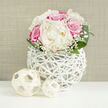 A rounded bouquet of white and pink blooms stands poised on a bright white tabletop, radiating a sense of calm celebration that feels at home in Epping's quiet residential streets and wedding venues. At its core, large creamy white peonies open generously, their many layers of velvety petals forming full, cupped blossoms that suggest softness and gentle luxury. Woven among them are rich fuchsia-pink roses, the sort of rose that draws the eye from across a room, with deep, saturated petals that subtly curl at the tips. Between these focal flowers, fine sprays of baby's breath thread through the design, each tiny white floret catching the light and creating a delicate, cloud-like veil that breaks up the stronger blocks of colour. Broad, dark green leaves curve neatly around the base, their slightly glossy finish grounding the bouquet and providing a natural frame. The flowers sit inside a distinctive white vase crafted from interlaced strands, like painted wicker or rattan, forming an airy, basket-like structure that feels both contemporary and rooted in nature. Through the open spaces of this lattice, the stems and inner shadows are just visible, adding depth and intrigue. In front of the vase lie two small woven balls in a similar pale, natural tone, one slightly larger than the other, echoing the same textural theme and drawing the eye along the surface. The entire arrangement is set against a softly textured beige backdrop, reminiscent of linen or a painted plaster wall, which lets the colours glow without distraction. This display would suit a registry ceremony near Epping High Street, a relaxed reception in Theydon Bois, or an elegant anniversary dinner delivered fresh by a local florist.