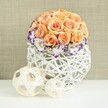 A refined floral display stands poised on a white tabletop, evoking the quiet sophistication of a carefully laid wedding table at an Epping venue. The focal point is a generously full, spherical dome of roses in soft peach and apricot hues. Each rose is at its peak, the petals lush and layered, creating a smooth, almost seamless curve of colour. The apricot tones vary gently from bloom to bloom, giving the dome a natural depth and subtle movement, as if lit by soft candlelight. Nestled around the lower edge of this rose globe is a ring of elegant lisianthus, predominantly white but intricately edged in rich violet or purple, like fine china painted by hand. Their frilled petals bring a gentle, romantic flutter to the arrangement. Scattered between the lisianthus are delicate sprays of white baby's breath, their tiny blossoms catching the light and forming a gossamer-like frame at the base of the dome. All the flowers are held in a distinctive, spherical white container woven from rattan or a similar material. Its open lattice structure allows glimpses of stems and shadows inside, adding texture and an airy, contemporary feel. The container's bright white tone contrasts cleanly with the warmer flowers and the soft beige fabric backdrop, which has a subtle, linen-like weave. In front of the centerpiece sit two decorative woven spheres in natural, pale wood tones, one larger and one smaller, echoing the container's craftsmanship and adding a sculptural flourish. The overall impression is of calm romance and understated luxury, a design that would feel perfectly at home at a bridal shower, anniversary dinner, or intimate celebration in an Epping home or boutique restaurant.