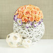 This image presents a lovingly assembled bouquet that reads both elegant and approachable: a compact dome of about twenty-four to thirty peach-apricot roses, each bloom layered with soft, satiny petals that catch the light and suggest a gentle rose scent. Around their lower edge, white Lisianthus flowers open like porcelain cups, each petal edged with a fine, dramatic sweep of deep purple that creates a vivid rim against the warm roses. Scattered throughout are tiny, cloud-like sprigs of baby's breath (Gypsophila), which give the massing a buoyant, garden-fresh lightness. The entire cluster is seated in a rounded white woven vase, its open lattice of reed or wicker lending a handmade, almost airy architecture that lets shadows fall playfully through the gaps. To the left of the arrangement two woven wooden spheres - one slightly larger than the other - rest on the table, their pale, natural fibres offering a tactile counterpoint and a hint of organic styling. A soft, linen-hued backdrop fades gently to keep attention on the flowers, while the soft daylight suggests a calm morning on a local high street or at home. The arrangement feels well-suited as a thoughtful gift for anniversaries, birthdays, or to lift a living room with quiet charm - a sophisticated but warm statement that balances texture, colour and light with a refined, contemporary eye.