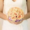 A softly lit, chest-level close-up shows a bride in Epping cradling a round bridal bouquet, the flowers glowing gently against the creamy white of her lace wedding gown. The bodice of the dress is detailed with fine, traditional lacework and a subtle V-shaped neckline, while the skirt falls in smooth, elegant folds that frame her hands. At the heart of the image is the bouquet itself: a lush dome of tightly clustered roses in tender shades of peach, apricot, and blush pink, each bloom layered with velvety petals that curl inward like whispered secrets. The roses form a warm, romantic core, their colours reminiscent of a soft Epping Forest sunrise on a clear spring morning. Circling beneath this rose canopy is a defining ring of deep purple blooms, likely lisianthus, their ruffled petals tipped and washed with white so that each flower appears to be brushed with light at the edges. This purple-and-cream border adds depth and a gentle drama, making the pastel roses seem to float above it. Threaded through and around the base are tiny white clusters of baby's breath, adding an airy, cloudlike softness. The bride's hands, with light, understated manicure and a hint of shimmer on one thumb, clasp the stems with quiet assurance. There is no visible ribbon, just the impression of a carefully bound, hand-tied arrangement chosen with intention. The overall mood is one of poised calm and intimate joy, as if captured just outside a church on Epping High Street or before stepping into a local venue, the bouquet ready to be remembered in both photographs and memory as a symbol of the day's love and elegance.