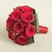 A finely arranged bridal bouquet, echoing the natural romanticism found through the heart of Epping, features a compact cluster of eight to ten deep red roses, each bloom opening to reveal velvety, richly coloured petals at peak freshness. Among the roses, small globes of red hypericum berries catch the light, their smooth surfaces supplying playful contrast and subtle festive vibrancy. Encircling the bouquet is a collection of broad, dark green leaves, carefully wrapped for both structure and flair, recalling the lush borders of Epping's famed cricket fields. The stems are gathered tightly and bound with a lustrous crimson satin ribbon, creating an elegant and comfortable handle-perfect for moments of celebration. The entire arrangement rests on a neutral-toned, textured linen fabric, which brings both warmth and sophistication while allowing every shade of red and green to stand out. Under gentle, diffuse lighting, the interplay of textures-silky petals, glossy berries, and soft ribbon-delivers a sensual delight. This bouquet, favoured by many Epping couples for weddings and special occasions, embodies deep love and devotion, offering a glimpse into the thoughtful workmanship that local florists bring to every handcrafted design.