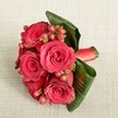 A richly romantic hand-tied bouquet of deep red roses and berries rests on a soft, light beige textured fabric, captured from a slightly elevated angle so every detail of the floristry is visible. Around five or six velvety roses in full bloom form the heart of the design, their petals layered in spirals of crimson and scarlet that seem to glow against the neutral background. Between the roses, clusters of small, rounded Hypericum berries in shades of red and reddish-orange add sparkle and texture, like little jewels nesting among the blooms. Broad, dark green tropical leaves are curved and folded with care, wrapping partway around the bouquet to create a natural collar that both frames and supports the flowers. The leaves show gentle veining and a subtle sheen, giving the arrangement structure and depth. Near the base, a rich red satin ribbon is tightly wound around the stems, its smooth surface catching the light and echoing the colour of the roses. The lighting is soft and even, with no harsh shadows, emphasising the luxurious textures of petal, berry, and foliage. The bouquet feels intimate and elegant-perfect as a boutonnière-style accent for a formal event in Epping, perhaps at a celebration near Epping Forest or along the High Street, or as a small romantic gift delivered locally. The overall impression is of a carefully crafted floral piece that radiates warmth, passion, and understated sophistication, suitable for weddings, anniversaries, or any special moment where a touch of classic red romance is desired.