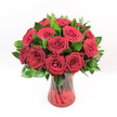 A studio-style photograph shows a classic, romantic bouquet of deep red roses gathered in a ruby-red glass vase, set against a perfectly clean white background that makes every petal stand out. Around a dozen or slightly more velvety crimson blooms are clustered tightly together, each rose at that ideal stage of opening where the centre is still gently furled, yet the outer petals curl outward in soft spirals. The petals have the richness of red wine and the texture of brushed velvet, giving the whole arrangement an indulgent, almost cinematic feel. Between the roses, glossy dark green foliage, likely Ruscus, peeks through, its pointed leaves catching small highlights of light and framing the flowers with a natural, elegant contrast. The foliage curves around the base of the arrangement, helping the bouquet form a full, rounded dome that feels carefully balanced yet effortlessly lush. The roses sit in a translucent, fluted red glass vase with a subtle ribbed or swirling texture that glimmers under the bright, even lighting, a detail that would feel smooth yet finely ridged to the touch. Through the coloured glass, the darker vertical lines of the stems can be seen, criss-crossing and overlapping as they descend into the water, adding depth and a sense of real, fresh life. The image feels like a timeless symbol of passion and devotion, the kind of bouquet you might send to a loved one in Epping for a milestone anniversary, a Valentine's Day surprise, or a dramatic romantic gesture that lights up a home near Epping Forest or along High Street with warmth, love, and uncomplicated elegance.