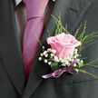 Pinned securely to a charcoal-grey jacket, this pink rose boutonniere appears like a small, intimate garden carried close to the heart. The central bloom, a soft, shell-pink rose, is fully formed yet still tender at the edges, with petals that curl outwards in smooth, velvety layers. It has that freshly opened look you might see on a mild spring morning walking through the green spaces around Epping, where the air is cool and moist. Surrounding the rose, light, airy sprigs of gypsophila create a cloud of bright white dots, each tiny blossom adding a sense of innocence and celebration. Wispy, needle-like greenery fans out behind and alongside the rose, adding structure, depth, and a gentle contrast, reminiscent of the foliage you might find bordering paths near Epping Forest. The stems are bound with a lilac satin ribbon, carefully wrapped and tied so that the finish is sleek and considered, the ribbon catching a soft reflection of light similar to polished silk. The overall palette-pink, white, deep green, and a hint of lilac-is calm and romantic, suited perfectly to weddings, civil ceremonies, and special events in and around Epping. You can imagine the subtle fragrance: a sweet rose scent combined with the fresh, almost resinous note of the greenery, grounding the sweetness with something quietly natural. Crafted by an experienced local florist, this boutonniere feels both timeless and personal, the sort of floral detail chosen thoughtfully for a groom, best man, or loved one to wear proudly as they share in a day of joy and connection.