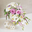 A romantic floral basket arrangement sits poised on a glossy white surface, glowing as if caught in soft morning light. At its heart, fully opened pink roses form lush, rounded blooms, their layered petals dense and velvety, suggesting quiet luxury and heartfelt affection. They are surrounded by cascades of white Phalaenopsis orchids, their smooth, wing-like petals and subtle golden throats giving the arrangement a refined, modern edge often seen in elegant Epping wedding settings. White lisianthus blooms and green-tipped buds, some kissed with gentle lavender at the ruffled edges, nestle between the roses and orchids, helping the eye travel in a sweeping arc from one side of the basket to the other. Wisps of gypsophila float like tiny stars among the larger flowers, bringing airy lightness and a sense of movement, as though a breeze from Epping Forest had just brushed past. Tall, slender stems of deep purple veronica emerge from the mass of petals, adding height and a hint of drama to the otherwise soft, cascading form. The flowers are arranged in a shallow white wicker basket with a detailed, cross-weave pattern that feels handcrafted and homely, perfect for gracing a wedding table in Epping's local hotels or countryside barns. Two pale rattan decorative balls rest in front, one medium and one small, their open lattice pattern mirrored in the basket's texture. A muted taupe fabric backdrop completes the scene, ensuring all attention stays on the bouquet's blend of whites, pinks, lilacs, and fresh greens, evoking the freshness of a spring celebration with local flower delivery just moments away.