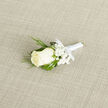 Resting on a pale beige woven surface, this boutonniere is an exercise in gentle craftsmanship: a single ivory rosebud anchors the composition, its petals folded in a quiet spiral that reads both classic and intimate. Tiny sprays of baby's breath form a soft, cloudlike frame, their pure white dots drifting around the rose and lightening the arrangement. The supporting greenery is thoughtfully varied - narrow, feathery twigs that evoke the texture of evergreen or cedar sit alongside broader, glossy deep-green leaves that lend structure and contrast. A smooth white satin ribbon binds the stems with precision, finished in a modest bow that keeps the silhouette neat and wearable. Under diffused daylight the cream of the rose and the green tones feel fresh and calm, and the overall impression is of a boutonniere prepared with steady hands and an eye for balance. It's the kind of accessory chosen for a ceremony on a crisp spring morning, a small emblem of purity and new beginnings to be pinned to a lapel and carried throughout vows and photographs. Imagine this piece delivered for a wedding reception in the Epping area, arriving with that faint, almost powdery floral scent and the reassuring professionalism of a local florist who knows how details matter on the big day. The neutral background enhances the textures and allows every petal, frond, and ribbon fold to be read clearly, an intimate composition meant to sit close to the heart.