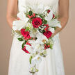 The image focuses closely on a bride holding a dramatic cascading bouquet, her arms and the bodice of her white, lace-detailed gown framing a luxurious mix of red and white flowers. At the centre, plush crimson roses, each bloom full and spiralled, create a passionate core that anchors the design with rich colour and sentiment. Nestled between them are pristine white lilies, opening wide like stars, their creamy petals and pale green throats giving the arrangement a refined, almost porcelain elegance. Flowing from the bouquet's base, delicate white Phalaenopsis orchids tumble gracefully downward, some flowers fully open, others still in bud, creating a soft waterfall effect that moves naturally with the bride's step. Wisps of white baby's breath weave gently through the composition, adding a light, cloud-like softness that contrasts beautifully with the glossy red hypericum-style berries, which dot the greenery like tiny, cheerful lanterns. A mix of slender fern fronds and broader green leaves curves around the flowers, highlighting their colours and giving the bouquet a fresh-from-the-studio vibrancy, reminiscent of the local flower stalls near Epping High Street. The softly blurred, neutral background keeps the viewer's attention firmly on the bouquet and gown, suggesting a quiet, intimate moment just before the walk down the aisle. Crafted with the eye of a seasoned Epping florist, the bouquet in this photograph feels both classic and contemporary, a red and white bridal statement perfect for couples marrying in local churches or rustic barns on the edge of Epping Forest, and ideal for inspiring wedding bouquet ideas in this area.