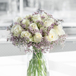 On a gleaming white tabletop, a clear cylindrical glass vase holds a lush, dome-shaped bouquet of creamy white roses and airy lavender filler flowers, the sort of arrangement you might find gracing a calm sitting room in Epping during a quiet afternoon. The roses are abundant and meticulously arranged, their petals unfolding in soft, spiralled layers that range from tight, promising buds to fully open blooms with wide, cupped faces. Subtle hints of pale green on the outer petals add a cool freshness, echoing the soft greens of nearby Epping Forest. Nestled between these roses are fine stems of purple statice, each carrying dozens of tiny, clustered florets that form a delicate, cloud-like frame around the central blooms. The contrast between the velvety rose petals and the slightly papery texture of the statice creates a pleasing interplay that can almost be felt through description alone. The clear water in the vase reveals a bundle of straight, neatly trimmed green stems, aligned with the precision of an experienced florist. Soft natural light floods the scene, casting gentle highlights and faint shadows that lend the composition depth and calm, while the background dissolves into an unobtrusive blur of pale grey and white. The mood is tender, reflective, and respectful, making this bouquet a thoughtful choice for sympathy flowers in Epping-a quiet companion on a mantelpiece, beside a condolence card, or in a small chapel alcove, offering a serene presence when comforting beauty is most needed.