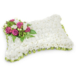 A soft, rectangular pillow-shaped tribute made almost entirely of tiny, pristine white chrysanthemums forms the heart of this memorial arrangement. The densely packed white blooms create a seamless, plush surface that reads like a textile-gentle, matte, and quiet-inviting a viewer to imagine the faint, clean scent of chrysanthemums and the calm they bring. In the upper-left corner a concentrated spray of deep magenta-fuchsia roses provides a bold, heartfelt focal point; their velvety petals open in layered spirals, offering warmth against the cool white field. Interspersed among the roses are small clusters of delicate white freesias or similar tiny white buds, their presence adding a soft, honeyed fragrance and a delicate punctuation of innocence beside the richer roses. Lush green foliage, including slender fern fronds and glossy leaves, fans out beneath and around the accent cluster, giving the composition a natural framework and a sense of graceful movement. A narrow, light green satin ribbon-pleated or looped carefully along the pillow's edge-peeks out beneath the chrysanthemums, introducing a whisper of colour and a finished, ceremonial border that defines the shape. The arrangement is photographed against a crisp white background with gentle shadowing under the pillow, which highlights its three-dimensional sculptural quality. This classic sympathy pillow, crafted with attentive hands and subtle scent, conveys rest, peace, and enduring remembrance-a tactile, floral expression intended to comfort mourners in services or graveside farewells in the Epping area, near the quiet lanes that lead toward Epping Forest.