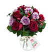 A lush, hand-arranged bouquet of deep crimson and soft lavender roses sits in a rounded, antique-style mercury glass vase, the colours vivid against a clean white background. The roses-rich burgundy and true red alongside gentle lilac and dusty purple blooms-are fully open, their velvety petals unfurling in layered concentric spirals that invite the fingertips to linger. Dark glossy greenery, likely Ruscus or Salal, threads through the composition, its cool, leathery leaves setting off the roses' warmth and giving the arrangement a dense, sculpted silhouette. Scattered among the roses, small red-orange rose hips or hypericum-like berries catch the eye with bright pops of seasonal colour and a slightly matte, rustic texture that contrasts with the smooth petals. The vase itself is distinctive: a bulbous base tapering to a narrower neck, finished in a mottled silvered mercury-glass effect that reflects soft highlights and lends a quiet, vintage glamour. A simple length of natural raffia or twine is tied around the neck, from which a small rectangular white gift tag hangs, showing a delicate stylized rose illustration and a short handwritten note in green and pink inks. Soft studio light washes the scene, accentuating the depth of the reds and the coolness of the lavenders while casting gentle shadows across the foliage. This composition reads as romantic and considered-perfect for an anniversary, an intimate celebration, or as a thoughtful gift to brighten a home in Epping or for local delivery to nearby cottages by Epping Forest.