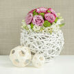 On a smooth, reflective white tabletop, a sophisticated floral centerpiece takes pride of place, the kind you might find gracing a wedding table at a country venue just outside Epping. At its heart sits a round, sculptural vase crafted from white-painted twigs or wicker branches, interwoven into a hollow sphere that feels both rustic and contemporary. Through its open framework, you catch glimpses of stems and shadows, adding depth and an airy lightness. Rising from this unique vessel is a carefully shaped dome of blooms: soft pastel pink roses, their petals full and velvety, mingle with a few deeper-edged buds that introduce a subtle blush tone. Nestled among them, vivid lime green button chrysanthemums create a lively mosaic of tiny, rounded pom-poms, bringing energy and contrast to the otherwise gentle palette. Encircling the base, a scattering of delicate white flowers with creamy centers - perhaps small lisianthus or spray blossoms - form a soft collar that blurs the line between the tight bouquet and the woven vase. To the side, on the same white surface, sit two decorative wooden spheres in pale, natural tones, their open lattice design echoing the vase's pattern and reinforcing the theme of organic craftsmanship. Behind everything, a warm taupe, fabric-like background remains slightly out of focus, ensuring that the roses and chrysanthemums remain the focal point. The scene feels serene and polished, ideal for a couple seeking a fresh, modern floral design for their wedding reception, whether they are celebrating in central Epping, Theydon Bois, or a nearby countryside barn.