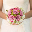 Held closely against the intricate lace of a bridal gown, this round hand-tied bouquet captures the tender, anticipatory hush just before the ceremony, the sort of calm you might feel standing in a quiet corner of Stonards Hill park. At its heart, full-headed roses in a soft blend of lavender and dusty pink unfold in perfectly layered petals, velvety and resilient to the touch, each bloom echoing a promise made. Nestled among them, zesty lime-green chrysanthemums cluster like little pompons, lending a fresh, playful contrast that lifts the entire arrangement into something distinctly contemporary while remaining timelessly romantic. Surrounding this central harmony is a delicate collar of creamy white flowers and unopened buds, their tapering shapes and smooth petals forming a gentle, lace-like rim that mirrors the bridal dress beneath. The light catches on every curve of petal and hint of green, creating subtle shadows that give the bouquet a feeling of depth and quiet movement. Imagine the fragrance: soft rose perfume mingled with cool, green notes, calming the bride's hands as she holds it close on the way to the aisle. Crafted with the practiced eye of a local Epping florist, each stem is positioned for balance and longevity so the bouquet appears as fresh at the reception near Bell Common as it did in the morning light. The result is a floral embrace that speaks of joy, hope, and new beginnings, perfectly suited to a heartfelt wedding day in and around Epping.