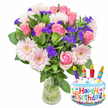 A joyful birthday arrangement photographed against a clean white backdrop: a lush hand-gathered bouquet in a clear glass vase sits slightly left of centre, the water and green stems visible through the vase's gently widened base. At the heart of the composition are numerous soft pink roses, their velvety petals opening from pale blush to a deeper rosy tone, giving a romantic, warm core to the display. Interwoven among the roses are light pink Gerbera daisies with broad, layered petals and pale yellow-to-soft-pink centres that read like smiling faces. Bright, clustered sprigs of vivid purple filler flowers - likely statice or limonium - are threaded through the arrangement, their crisp, papery texture creating contrast and depth against the silky rose petals. Rich, glossy green foliage frames the blooms, leaves of varying shapes and sizes building a lush backdrop that grounds the colour palette. To the lower right, a cheerful cake-shaped metallic Mylar balloon complements the bouquet: the balloon's white 'frosting' is edged with a wavy blue piping, scattered with red, yellow, blue and green sprinkles, and topped with four striped candles - purple, red, orange and blue - each bearing a tiny yellow flame. Across the front, Happy Birthday is written in playful multi-coloured letters, and the small 'KALEIDOSCOPE' brand mark sits at the balloon's base. Soft, natural light suggests morning delivery, and the overall mood is bright, celebratory, and thoughtful - an ideal gift for a birthday in Epping, whether collected from a High Street visit or sent across town for same-day delivery.