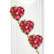 Three sumptuous hearts made entirely of roses form a cascading tribute, aligned in a gentle diagonal as though drifting down a quiet, whitewashed wall in a peaceful Epping farewell setting. Each heart is densely woven from fresh roses in deep, velvety crimson, vivid fuchsia pink, and soft blush tones, their petals overlapping to create a richly padded, almost quilted effect. The variation in bloom stages-from tightly furled buds to fully open flowers-adds layers of interest and a sense of unfolding emotion. Outlining every heart, fine strands of lush green ivy trace the curves with care, the small leaves adding an organic rhythm that softens the strong heart shapes. These ivy tendrils extend downwards to link one heart with the next, creating a visible chain of connection that can suggest family ties, enduring partnership, or generations of shared memories. The bright white background behaves almost like a gallery wall, reflecting light back onto the roses so their reds and pinks shine clearly, while faint shadows give the hearts depth and presence. The mood is romantic yet respectful, perfectly suited to a funeral or memorial tribute for someone cherished in the Epping area. The composition reflects the thoughtful hands of an experienced florist, using classic roses and natural greenery to convey love, remembrance, and a gentle sense of continuity, like a final message spoken softly in flowers.
