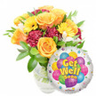 A bright, uplifting 'Get Well Soon' gift ensemble photographed against a clean white backdrop: to the left a generous, classical bouquet sits in a clear glass vase with stems and water visible, and to the lower right a shiny, round silver Mylar balloon overlaps the blooms. The bouquet is built around several large, opening roses in warm orange and deep yellow tones whose velvety petals spiral softly outward, giving a comforting, citrus-warm scent. Sunny yellow gerbera daisies punctuate the arrangement with broad, cheerful faces, while deep burgundy-to-maroon carnations add ruffled texture and a rich counterpoint. Sprays of small white alstroemeria (Peruvian lilies) peek through the massed flowers, introducing delicate, airy accents and a faint sweet perfume. Abundant green foliage - glossy leaves of varied shapes and lengths - frames every bloom, lending freshness and a lively silhouette that reads as both natural and carefully shaped by a florist's hand. The round foil balloon has a reflective silver field decorated with a playful cluster of cartoon-style balloons in blues, purples, pinks, greens and yellows, some patterned with stripes, polka dots and checks; bold yellow-and-blue lettering in the centre proclaims Get Well Soon, and tiny starburst motifs ring the white border while a small KALEIDOSCOPE logo sits at the bottom edge. Bathed in soft daylight that highlights petals and metallic sheen, this arrangement radiates hope and gentle cheer - ideal for sending to a friend or neighbour in Epping town centre or for collection at a local market, a thoughtful companion for recovery and rest.