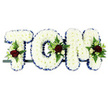 This image shows a traditional funeral name tribute spelling TOM in large, three-dimensional letters entirely covered with white chrysanthemums. The flowers are tightly clustered so that they form a smooth, padded surface, giving each letter a soft, cloud-like appearance on the clean white background. A royal blue ribbon trims the outer edge of every letter, neatly pleated and gently shimmering, which outlines the word clearly and adds a touch of solemn colour well-suited to a memorial service in Epping. At key points on the letters-near the upright of the T, the curve of the O, and the upper arm of the M-small, carefully placed sprays of feature flowers break the white expanse. These include rich, deep red roses open at their centres, smaller white or cream spray roses, and slender, pure white blooms reminiscent of freesia, all framed by lush green foliage and feathery ferns that add texture and a natural, garden-like feel. The letters rest along a narrow, dark frame that runs beneath them, keeping the tribute straight and stable, suitable for placement by the coffin, at the front of a chapel, or in a quieter corner of a wake at a local venue. The colour combination of white, blue, and red suggests purity, tranquility, and devoted love, while the named design gives the piece an intimate, personalised character. It feels like a gentle, dignified message in flowers for Tom, reflecting both the sorrow of loss and the tenderness of cherished memories shared by family and friends across the Epping community.