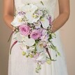 Amid the tranquil elegance of an Epping wedding, a stunning bridal bouquet commands the viewer's eye-cradled gently by a bride whose white gown's lace details shimmer in the soft afternoon light. This breathtaking arrangement, crafted in a luxurious cascading waterfall style, features pristine white Phalaenopsis orchids as its radiant centerpiece, their broad petals encircling vivid yellow-gold hearts. Surrounding these are lush clusters of blush pink and lavender roses, each bloom a tender testament to local romance and handcrafted expertise from Flowers Epping. Wisps of baby's breath meander through the bouquet, infusing it with lightness, while smaller lavender-toned blossoms evoke the gentle wildflowers found outside Epping's town center. The fluid design unfolds downward, with more delicate orchids, slender buds, and distinctive magenta amaranthus providing tactile drama and textural intrigue. Subtle greenery and the curve of each leaf add freshness, echoing the natural beauty seen throughout Epping's nearby woods and parks. The color palette-soft whites, romantic pinks, serene purples-creates an impression of timeless grace, ideal for spring or summer countryside weddings. The image captures more than just a floral creation; it reflects the anticipation, love, and promise of the day-each petal and stem thoughtfully placed to inspire lasting memories for any local bride planning her ceremony in this picturesque corner of Essex.