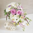 A sophisticated floral display overflows from a round white wicker basket, styled as a low wedding table arrangement that would sit beautifully at a reception in Epping or a nearby village. The bouquet is dominated by romantic shades of pink and white, with several large blush-pink roses fully opened at the centre, their petals forming tight yet generous spirals that suggest both luxury and warmth. Above and around them, sleek stems of white Phalaenopsis orchids drift gracefully, their wide, satin-like petals and golden-yellow centres lending a refined, almost sculptural quality to the design, perfect for an intimate celebration meal. Tucked between these feature blooms are frilled white Lisianthus flowers, some touched with a hint of lilac along the edge of each petal, adding a delicate cool tone that enriches the composition. Branching from the sides, soft pink stock flowers extend in airy clusters, providing gentle height, fragrance, and a slightly wild garden feel reminiscent of borders around homes near Epping High Road. Fine sprays of white baby's breath thread through every layer, filling gaps with a gossamer texture that makes the entire arrangement feel light and cloud-like despite its fullness. Glossy green foliage and slender shoots emerge between the flowers and slip gracefully over the edge of the basket, keeping the look fresh and natural rather than overly formal. The basket sits on a bright white surface, its tightly woven texture mirrored by two small decorative spheres crafted from interlaced pale wicker placed close by. Behind, a soft beige, linen-like backdrop with a gentle weave keeps the scene calm and neutral, allowing the pastel tones and florist artistry to shine, ideal for describing a fresh, local wedding table arrangement in the Epping area.