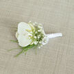 An elegant white orchid boutonnière is displayed on a pale, sandy-beige fabric that looks like fine linen, its subtle weave catching the light in a gentle, understated way. At the heart of the design is a single, immaculate white orchid bloom, its petals broad, velvety, and unblemished, opening with confident grace. Inside the orchid's centre, a whisper of creamy yellow and soft butter highlights the inner details, adding warmth and dimension to the icy-white outer petals. Encircling this central bloom are airy clusters of white baby's breath, each spray of tiny flowers forming a delicate, cloud-like border that enhances the orchid's larger, sculptural form. Slim blades of fresh, bright green foliage thread through the arrangement, their clean lines and fine tips suggesting ornamental grass or slender fern fronds, echoing the natural greenery you might see around Bell Common or along the edges of Epping Forest. The stems beneath are meticulously gathered and wrapped in smooth white satin ribbon, creating a neat, cylindrical handle that would sit comfortably on a suit jacket. Near the top, a small satin bow is tied with precision, introducing a final, classic flourish that speaks of an experienced florist's touch. The boutonnière lies at a gentle angle, the soft, even lighting accentuating its textures-the satin's sheen, the orchid's satiny petals, the feathery gypsophila, and the woven background. The mood is one of polished simplicity and quiet romance, perfectly suited to a wedding ceremony at a local Epping venue, where this piece might adorn the lapel of a groom, best man, or cherished family member, adding a hint of natural elegance to an already special day.