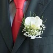A close-up photograph captures the upper torso of a person dressed for a special celebration in Epping, their formal wear brought to life by a striking orchid buttonhole. A deep charcoal or black suit jacket fills most of the frame, its lapel cleanly structured and sharply pressed, suggesting a groom or prom guest standing ready just before the event. Against the dark fabric sits a pristine white Phalaenopsis orchid, its broad, velvety petals fanning out like folded silk, with a soft yellow-cream throat that seems to glow gently under natural light. Clustered around the orchid is a halo of tiny white baby's breath blossoms, each delicate flower creating a misty, cloud-like texture that softens the crisp lines of the suit. Slim, grass-like green foliage arcs gracefully from behind the blooms, adding freshness and movement, as if echoing the greenery of nearby Epping Forest on a spring morning. The stems are neatly wrapped and finished with a fine white sheer ribbon, tied into a small, perfectly formed bow that lies flat against the lapel, giving a polished, refined look. Just beside the boutonniere, a lustrous red silk tie sweeps down the chest, rich and bold against a subtly patterned, light dress shirt, hinting at chequered detailing that adds quiet character. The tie's sheen catches the light, bringing warmth and intensity to the composition, and suggesting romance, joy, and festive energy. This intimate portrait of formal attire feels like a moment paused outside a registry office or local Epping church, focusing on the florist's careful craftsmanship and the quiet significance of wedding-day or black-tie details worn close to the heart.