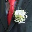 The image shows a refined wedding boutonnière resting in the breast pocket of a dark, expertly cut suit, the sort you might see at an elegant ceremony near Epping Forest or a classic venue off Theydon Road. The focal flower is a single white orchid, its petals broad and silky, unfolding in a calm, open shape that feels serene and composed. A soft, creamy sheen plays across the bloom, while at its centre a barely-there flush of warm gold hints at hidden depth and emotion. The orchid is cocooned by a halo of baby's breath, each minute white blossom distinct yet collectively forming a cloud-like softness, as if a light fog has settled just above the pocket. Fine green foliage threads through and around the composition, adding a fresh, natural rhythm that suggests movement without disturbing the design's poise. At the base, a slim white ribbon gathers the stems, its tidy bow and smooth texture revealing the careful craftsmanship of a local Epping florist who understands bridal details and groom's finishing touches. The boutonnière is framed by the strong line of a charcoal lapel and the bold splash of a vivid red tie, the contrast making the white petals appear even more radiant. There is an air of celebration and anticipation in this small floral accent, as if the wearer is moments away from walking into a ceremony at a nearby church or registry office, carrying with them the quiet fragrance, timeless style, and heartfelt symbolism of a thoughtfully designed wedding flower.