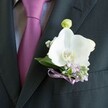 A close-up photograph reveals a meticulously tailored charcoal or deep black suit jacket, its left lapel sharply defined beneath soft, flattering indoor light, as a pristine white Phalaenopsis orchid boutonniere becomes the quiet star of the frame. The jacket fabric appears smooth and finely woven, sitting over a pale grey dress shirt with a subtle, almost herringbone texture, while a rich magenta or plum tie glows silkily at the centre, its knot sitting neat and confident at the collar. Pinned to the lapel, the orchid's broad, velvety petals open like porcelain wings, immaculate and gently curved, cradling a vivid yellow and soft orange centre that draws the eye with sculptural precision. Around this central bloom, tiny tufts of lilac or soft lavender flowers weave between airy sprigs of white gypsophila, or baby's breath, giving the arrangement a lace-like halo of texture and softness. Fresh green foliage peeks from behind, framing the flowers with a natural, polished edge, while a single pale green orchid bud stands just above the main bloom, suggesting new beginnings and quiet anticipation. The stems are wrapped and finished with a delicate satin ribbon in lavender or mauve, tied into a careful bow that echoes the tones of both the filler flowers and the nearby tie. The entire composition feels like a modern groom's boutonniere, the kind a couple in Epping might choose for a refined wedding at a local venue near Epping Forest, its understated elegance aligning perfectly with a day of promises, photographs, and gentle spring air. The image captures not only the floral details, but also the mood of calm, romantic preparation before a cherished ceremony begins.