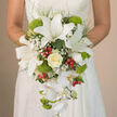 The image shows a bride standing quietly, her torso and hands in focus as she holds a cascading bridal bouquet that seems to flow like a white waterfall over the front of her lace gown. The dress itself is an off-white, with a subtle, intricate lace pattern that forms a delicate backdrop for the flowers. At the heart of the bouquet, large white oriental lilies open wide, each petal gently recurved and finely speckled with reddish-brown markings that draw attention to their prominent stamens. Just beneath the bride's fingers, a creamy ivory rose anchors the design, its velvety petals layered in a perfect spiral that speaks of timeless romance. Around it, tiny white gypsophila clouds soften the transitions between blooms, filling small gaps with a dreamy, airy texture. The bouquet tapers downward into a stream of white Phalaenopsis orchids, their arching stems and flat, elegant blooms giving the design a graceful, almost ethereal descent. Between the white flowers, vivid lime-green Green Trick dianthus or pompon chrysanthemums form small, rounded cushions of fuzzy texture that pop against the neutrals. Bright red hypericum berries are placed with care, adding lively spots of colour that hint at joy and celebration without overpowering the classic palette. Fine trailing greenery and darker foliage frame the cascade and give the bouquet a natural, garden-inspired feel, reminiscent of the lush greenery around Epping High Street or the edges of Epping Forest. The overall impression is of a handcrafted, thoughtfully balanced wedding bouquet, ideal for a bride who loves classic white flowers but wants a hint of personality and freshness in her wedding photos.